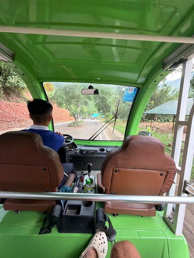A man is driving a green, open-air golf cart down a paved road in a rural area. The cart has two rows of brown leather seats. The driver, wearing a blue polo shirt and dark pants, is seated in the front left. The road is lined with trees and foliage, and a red, rocky embankment is visible to the left. A green roofed structure with rows of small plants is visible on the right side of the road. The weather appears to be overcast or rainy, as indicated by the droplets on the windshield. The scene is captured from the perspective of a passenger in the back seat. This location is identified as the city of Khanh Vinh, Vietnam.