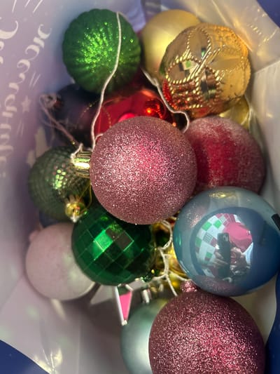 This is a close-up, high-angle shot of a collection of Christmas ornaments, primarily spherical, contained within a white bag that has a printed design featuring white cursive lettering and stars. The ornaments are of various colors and textures, including green, gold, red, pink, and blue. Some are smooth and shiny, while others are textured or covered in glitter. One blue ornament in the foreground reflects a person holding a smartphone, seemingly taking the photograph, and a pink cap. The lighting suggests an indoor setting. The image does not provide any visual information to determine the location of Arkhangelsk, Russia.