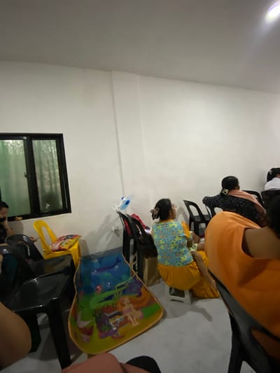 In a room with white walls and a grey floor, several people are seated, facing away from the camera. On the left, a black framed window with green tinted glass shows foliage outside. In front of the window, a yellow plastic chair holds a bag of chips. To the right of the yellow chair, a colorful child's bib or mat with animated characters lies on the floor. A person wearing a blue and yellow floral shirt and orange pants sits on a white stool. Another person in an orange shirt is partially visible on the right. Several black plastic chairs are scattered around the room. A small blue and white object, possibly a fan, is mounted on the wall to the right of the window. The scene appears to be indoors in San Jose del Monte, Philippines.