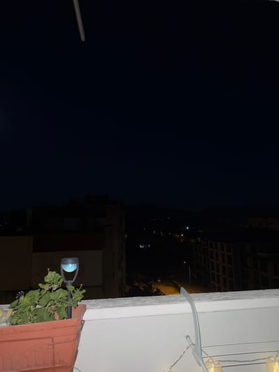 The image shows a nighttime view of a cityscape from a balcony. The scene is primarily dark, with the exception of some artificial lights. On the right side of the frame, a white wall is visible, with a decorative string of fairy lights draped over it. Below the fairy lights, a potted plant with green leaves sits on what appears to be a tiled surface, possibly part of the balcony's structure. Adjacent to the plant, a black outdoor light fixture is mounted on a pole, emitting a soft blue light.
The background reveals the dark sky and the illuminated windows of buildings in the distance. The perspective is from above, looking down and across at the urban environment. The time of day is clearly night, indicated by the darkness and the artificial lighting. There are no people or specific weather conditions visible. The overall impression is one of a quiet urban evening. The image appears to have been taken in Targovishte, Bulgaria.