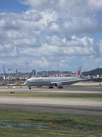 A wide-angle, eye-level, outdoor, daylight, full shot shows an Air China Boeing 787-9 Dreamliner on the tarmac of Shenzhen Bao'an International Airport in Bao'an District, China. The aircraft is predominantly white with red accents, featuring the Air China logo on its tail and "AIR CHINA" written along its fuselage. The plane is positioned slightly to the right of the frame, facing left and appearing to taxi or wait on the runway. In the background, a dense urban skyline with numerous buildings is visible, including construction cranes, suggesting ongoing development. The sky is filled with scattered, grey and white clouds against a bright blue sky. The foreground consists of airport tarmac and grass. No people are visible.
