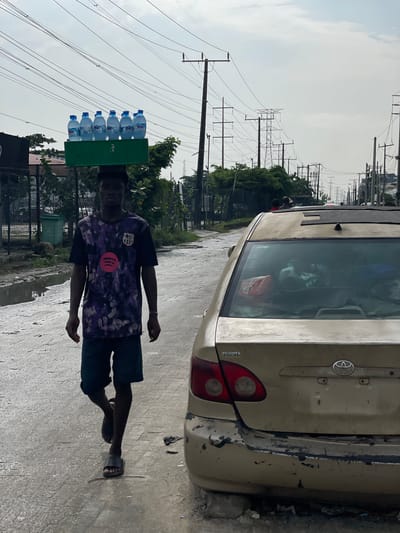 In Alaguntan, Nigeria, a young Black male walks along a wet, paved road, carrying a green crate of bottled water balanced on his head. He is wearing a dark t-shirt with a purple, black, and pink abstract design and black shorts. His feet are clad in black slides. To the right of the frame is the rear of a weathered, tan Toyota sedan, its bumper visibly damaged and scratched. The road appears to have standing water in puddles, suggesting recent rain or poor drainage. In the background, numerous electrical poles and power lines crisscross the sky, indicative of urban infrastructure. Trees and other vegetation are visible on the left side of the road, with a fence and some structures behind them. The sky is overcast.