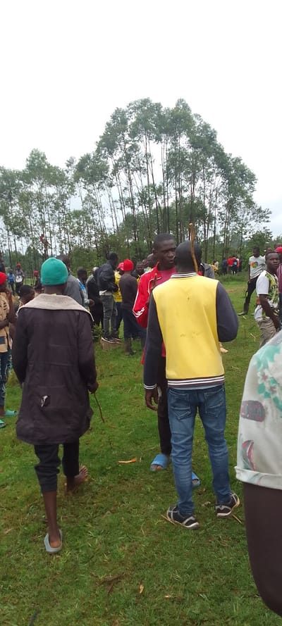 The image shows a group of people standing in a field, located near a forest in Lubao, Kenya (latitude: 0.378, longitude: 34.809). The individuals are gathered on a patch of green grass, with trees forming a backdrop behind them. Several people are wearing casual clothing, and one man in particular stands out wearing a bright yellow vest. He is speaking to another man, and they are both looking directly at the camera. Some people are standing around, while others appear to be walking away from the camera. The field is slightly uneven, and some small sticks and twigs are visible on the ground. There is a hint of overcast sky in the background, indicating the possibility of rain.