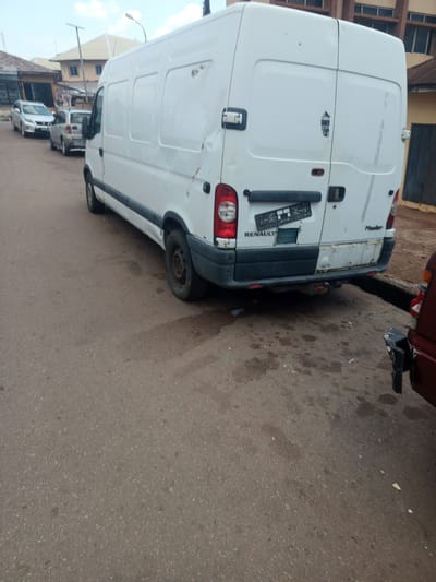 A white Renault Master van is parked on an asphalt road in front of buildings. The van is facing away from the camera, and its rear doors are closed. A license plate is attached to the rear of the van. To the left of the van, a silver SUV and a silver hatchback are parked. To the right of the van, the rear of a dark red vehicle is visible. The background shows buildings with windows and a street light. The ground is asphalt and appears damp in some areas.