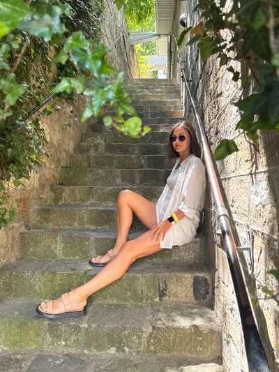 A young woman sits on stone stairs outdoors, leaning against a textured stone wall. She is wearing a white collared shirt with a subtle dark pattern, white shorts, and tan sandals. She has on round sunglasses and a yellow-strapped smartwatch on her left wrist. Ivy vines cascade down the stone wall to her left and on the right side of the frame. The stairs are made of large, rough-hewn stones, some of which are covered in moss. A metal handrail runs along the right side of the stairs. In the background, there is a glimpse of a covered walkway or structure with metal supports. The scene appears to be located in a sunny outdoor environment, possibly in the city of Abrau-Dyurso, Russia, known for its natural beauty and historical sites.
