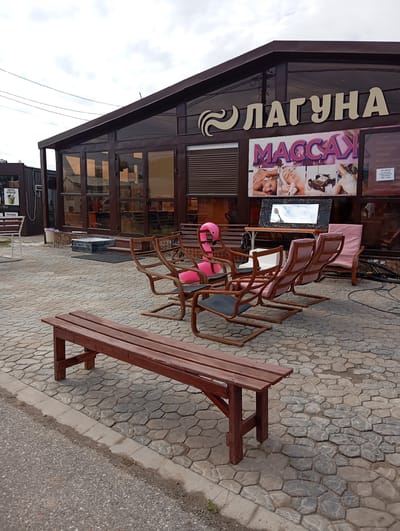 The image depicts the exterior of a commercial establishment in Sol-Iletsk, Russia, identified by the signage "ЛАГУНА" (LAGUNA) and "МАССАЖ" (MASSAGE). The building features large glass windows and a dark brown, angular roof. In front of the building, several chairs and benches are arranged on a stone-paved patio. A prominent pink inflatable flamingo sits on one of the chairs. Adjacent to the main building, another smaller structure with windows and signage is visible. The scene is outdoors, with a cloudy sky overhead. The overall impression is that of a relaxation or spa facility.