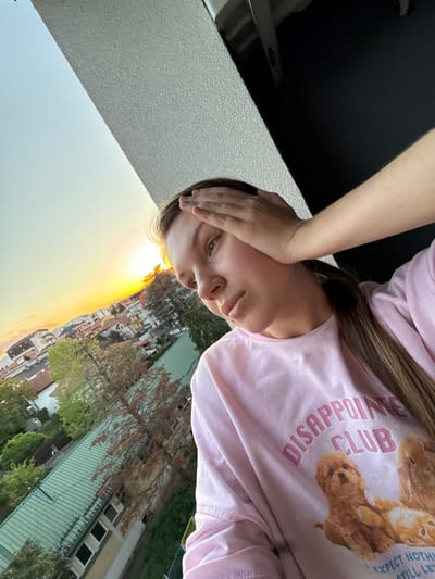 A selfie of a young woman wearing a pink long-sleeved shirt with a graphic of cartoon animals and the words "DISAPPOINTMENT CLUB", with her hand on her forehead. She has long brown hair. The background shows a cityscape at sunset. Based on the architectural style and foliage, it is likely that the location is in Vozdovac Urban Municipality, Serbia.