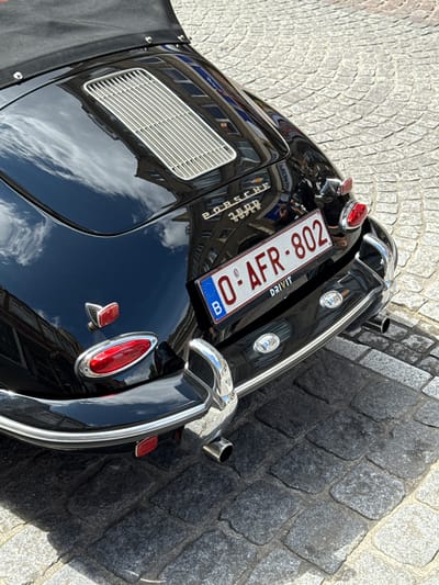 The image shows the rear of a black 1960s Porsche 356 Super convertible parked on a cobblestone street in Bruges, Belgium. The car has a black soft top partially visible. The license plate reads "0-AFR-802" and has a Belgian "B" sticker. The car's chrome trim, taillights, and exhaust pipes are visible. The street is paved with light grey and dark grey cobblestones, with shadows cast by the car and surrounding buildings. The reflection on the car's body shows buildings with windows. The overall scene is a street-level, outdoor shot taken during the daytime.