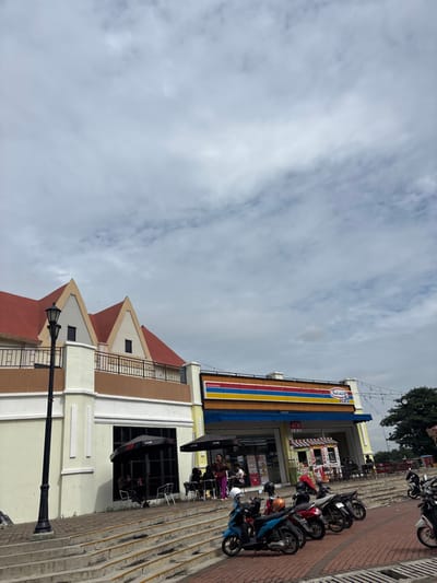 This media file captures a daytime street scene, likely in Bekasi Regency, Indonesia, under an overcast sky with light grey clouds. The primary subject is a multi-story building featuring a convenience store with a prominent blue awning and a large sign above it displaying horizontal stripes of blue, yellow, red, and white. Below the awning, an "ATM" sign is clearly visible. The building itself has a distinctive architectural style, characterized by light-colored walls (cream/white) with brown accents and multiple peaked gables on its roof.

In front of the convenience store, a paved area transitions into tiered concrete steps or seating, where numerous motorcycles are parked. Several people are visible in the scene; some are standing near the store's entrance, while others are seated at black outdoor tables and chairs, possibly enjoying a meal or resting. Black bases or stands, possibly for umbrellas, are marked with "LIVE" text. A classic black streetlamp stands in the foreground to the left. In the upper right background, some green trees are visible. The overall activity suggests a typical day with patrons visiting the convenience store and utilizing its outdoor amenities.