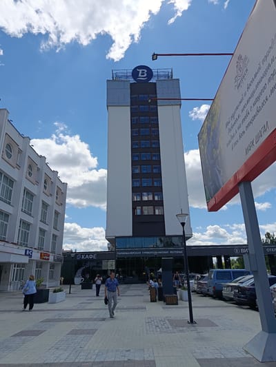 This is a vertical, eye-level, outdoor photograph taken on a sunny day with scattered clouds. The scene is a pedestrian plaza in front of a modern building complex. The primary subject is a tall, white and dark-grey building with many windows, topped with a large blue circle with a white "B" inside. To the left of this building is a shorter, white building with arched windows. In the foreground, people are walking across a paved plaza. Several shops and businesses are visible on the ground floor of the buildings, including "КАФЕ" (Cafe), "ПАРИКМАХЕРСКАЯ" (Hairdresser), "ТУРБЮРО" (Travel Agency), "БАНКОМАТ" (ATM), and "РЕСТОРАН" (Restaurant). A large billboard with text and an image is prominently displayed on the right side of the frame. The text on the billboard is in Russian and includes "ВИТЕБСК" (Vitebsk) and "МАРК ШАГАЛ" (Marc Chagall). The overall atmosphere is one of a bustling urban environment on a pleasant day.