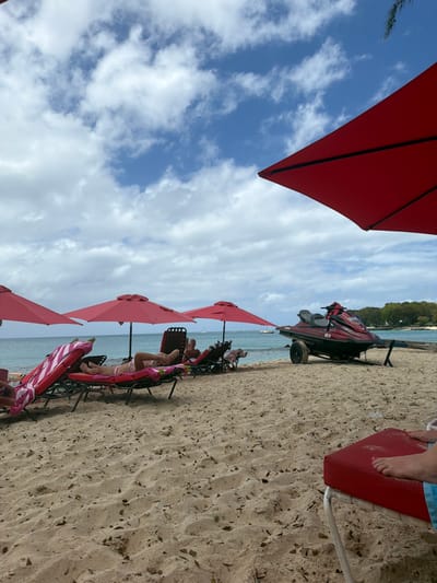 The scene depicts a sandy beach in Holetown, Barbados, under a partly cloudy sky with prominent white cumulus clouds and visible blue patches. In the lower right foreground, the edge of a red lounge chair cushion is visible, with a person's feet and lower legs, partially clad in blue swimwear, resting upon it.

In the midground, from left to center, three red beach umbrellas are deployed. Beneath these umbrellas, at least four individuals are present, reclining on lounge chairs. One person, wearing a pink swimsuit, lies prone on a red and white striped lounge chair. Another individual is partially visible reclining on a dark-framed chair further back. To the right of the lounging area, a red and black personal watercraft (jet ski) with the registration "488 H" is parked on the sand, mounted on a dark-colored two-wheeled trailer. The personal watercraft faces towards the ocean.

The background features the calm, turquoise water of the Caribbean Sea, dotted with several distant white boats or yachts. A lush, green-treed landmass forms the distant shoreline to the right. The beach sand is light brown with minor debris.
