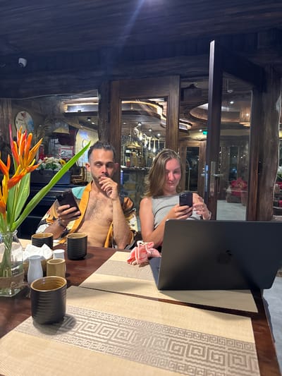 An exterior shot captures a scene with a man and a woman sitting at a table in what appears to be an outdoor seating area of a restaurant. The man, shirtless and wearing a patterned robe, is engrossed in his smartphone, holding it with his left hand. Beside him, a woman in a gray tank top is also looking at her smartphone. In front of them, a laptop is open on the table, its screen obscured. The table is set with placemats, cups, and a vase of vibrant orange and yellow flowers. The background reveals a rustic, wooden interior with exposed beams and traditional Vietnamese architectural elements, suggesting a location in Nam Hoa Lư Ward, Vietnam, possibly a restaurant or a guesthouse.