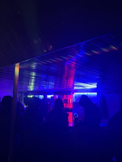 The image depicts a dimly lit indoor space, likely a bar or a club, illuminated by an array of blue and pink lights. The lighting reflects off a corrugated metal ceiling, creating streaks of color across its surface. In the foreground, several individuals are silhouetted against the light, their forms indistinct due to the low light conditions. They appear to be standing or milling about. In the background, a bar area is visible, with shelves lined with bottles and illuminated by red and white lights. A sign is also visible on the wall behind the bar. The environment suggests a social gathering or a nightlife establishment. There is no indication of the specific location being in Wumat, Nigeria.