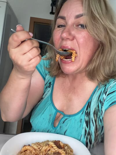 A middle-aged woman with blonde hair is captured in a close-up selfie as she eats pasta from a spoon. She is wearing a blue and black patterned top with a keyhole neckline. The background shows a glimpse of a kitchen or dining area, with a refrigerator on the left and a doorway to the right. The woman is leaning forward, bringing a spoonful of pasta towards her mouth. The lighting is bright, suggesting an indoor setting during the daytime. The location is Leningradskaya, Russia.
