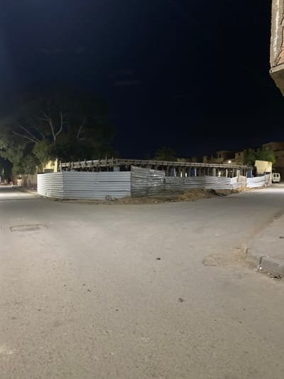 The image shows a street in Sidi Chahmi, Algeria, at night. The road is paved and appears to be somewhat worn. Along one side of the road, there is a series of vendor stalls or makeshift structures constructed from metal sheets. The stalls are aligned in a row, suggesting a market or commercial area. The sky is dark, and the presence of trees indicates the image was taken from a more vegetated part of town. A building is visible behind the stalls and the vendor stalls appear to be unoccupied.
