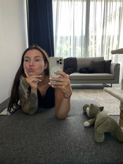 A woman is taking a selfie while lying on a grey carpet, with her head and upper body angled towards the camera. She has long, dark hair and is wearing a black top. Her right arm is bent, and her hand is resting on her chin. She is puckering her lips, creating a playful expression. Her left arm is partially visible and shows tattoos. She is holding a smartphone in her right hand, which is angled to capture the selfie.
In the background, a modern grey sofa with dark grey and striped cushions is visible, placed near large windows covered with sheer white blinds that allow natural light to filter through, suggesting it is daytime. A stuffed green animal, possibly a cat, is lying on a rug behind the sofa. The overall setting appears to be a well-lit indoor living space. No specific time of day or weather conditions can be determined beyond the presence of natural light. The location context of Kediri, Indonesia, is provided but not visually evident in the image. No visible text is discernible in the image.