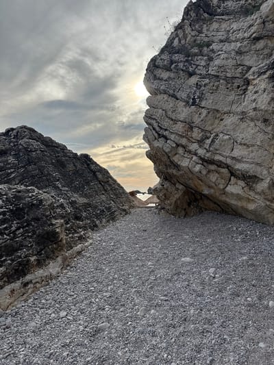 A coastal scene features a pebble beach leading between two large rock formations. The sky above is partly cloudy with the sun peeking through, casting a soft glow. In the distance, the sea is visible, and two thatched umbrellas suggest a nearby beach area. The foreground is dominated by the textured surfaces of the rocks and the multitude of grey pebbles covering the ground.