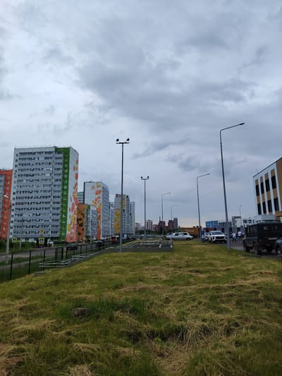 The image depicts an overcast day in a residential area of Kondratovo, Russia. A row of modern, multi-story apartment buildings with colorful facades line a street. In the foreground, a grassy slope with recently mown patches of hay leads down to a sidewalk and road. Several vehicles, including SUVs and sedans, are parked or driving on the street. A few pedestrians are visible on the sidewalk. Streetlights and some commercial buildings are also present in the scene. The overall impression is of a developing urban environment under a cloudy sky.