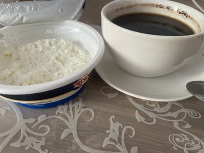 This image displays a close-up, eye-level view of a meal consisting of cottage cheese and coffee, likely on a table in Brovary, Ukraine. The cottage cheese is in a white plastic container with a blue and yellow label. The coffee is in a white ceramic cup with a saucer, and a silver spoon is visible on the saucer. The table surface has a decorative, light-colored pattern. The scene is static, with no visible people or actions occurring. The lighting appears to be natural, casting subtle shadows.