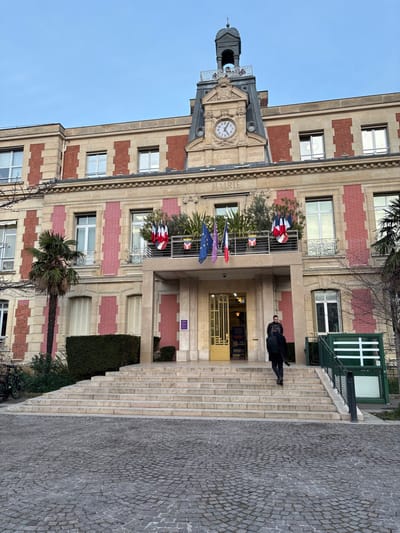 An external view of a grand building, identified as a "Mairie" (City Hall) in Alfortville, France, stands under a clear blue sky. The building features a distinctive facade of red brick and light-colored stone, with multiple windows, some fitted with white louvered shutters.
A prominent clock tower rises from the center of the building, crowned by a domed cupola. The clock face is visible, and beneath it, the national motto of France, "LIBERTÉ ÉGALITÉ FRATERNITÉ" (Liberty, Equality, Fraternity), is carved into the stone. Above the main entrance, the word "MAIRIE" is clearly legible.
The entrance is accessed by a wide, light-colored stone staircase. A single person, dressed in dark clothing, is seen walking up these steps towards the building. The main doorway is recessed and features intricate golden or bronze metalwork. Flanking the entrance on balconies are planters filled with green foliage. Several flags are displayed from poles on these balconies, including the French Tricolor and the flag of the European Union. Other national flags are also present, one distinctly appearing to be the flag of Portugal. Additionally, red, white, and blue decorative bunting is draped along the balconies.
To the right of the main staircase, a separate green structure with windows and a door, possibly an enclosed stairway or lift access, is visible. The ground in the foreground is paved with small, dark grey cobblestones. Vegetation includes a large palm tree to the left of the steps and various hedges and shrubs around the base of the building. The scene depicts daytime with bright, clear weather.