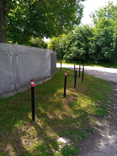 The image depicts an outdoor scene with a concrete wall on the left, a grassy area, and a dirt road on the right. A line of black bollards with red and white reflective bands is positioned along the edge of the grass, separating it from the road. Lush green trees are visible in the background, and the ground is illuminated by sunlight filtering through the foliage, casting shadows. There are no people visible in the image. The scene appears to be a pathway or a roadside area in a somewhat natural setting.
