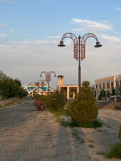 An outdoor scene in Taraz, Kazakhstan captures a pathway lined with trees and decorative benches leading towards a modern domed building. A prominent lamppost with ornate scrollwork and banners displaying Cyrillic script stands in the foreground. To the right, an arched building with Islamic architectural influences is visible. The pathway is paved with brickwork, and several large wagon wheels are integrated into the design of the benches. A lone individual is seated on one of these benches, facing away from the camera. The sky is partially clouded with a soft, warm light suggesting either early morning or late afternoon.
