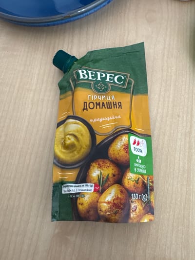 The image shows a package of mustard on a wooden surface. The mustard package is yellow and green with text in Ukrainian. It features a picture of potatoes and a swirl of mustard. There is also a blue plate in the upper left corner of the image. The scene is indoors, likely on a table. There is no indication of the city of Pilsen, Czechia.