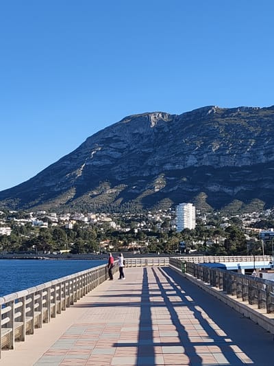 This vibrant image captures a sunny coastal scene in Dénia, Spain, showcasing a pier in the foreground, a town beside the sea in the midground, and a majestic mountain dominating the background under a clear sky.
In the foreground, a wide, paved pier extends from the bottom right towards the left, featuring a pattern of reddish-brown and lighter square tiles. A sturdy wooden railing lines the left edge of the pier, separating it from the water. Long, sharp shadows, cast by the railing and possibly other unseen structures, stretch diagonally across the pier, indicating a low sun angle, suggesting either early morning or late afternoon. Two individuals are standing near the railing on the left side of the pier, looking out towards the expansive view. One person is dressed in a red top and dark pants, while the other wears a white top and dark pants.
To the left of the pier, a calm, deep blue body of water, likely the Mediterranean Sea, stretches into the distance. Beyond the water, in the midground, lies a bustling coastal town. It is characterized by numerous white and light-colored buildings interspersed among abundant green trees and vegetation. A tall, modern white high-rise building stands out prominently within the town's landscape.
Dominating the background is a large, rugged mountain, showcasing distinct rock formations and slopes covered in a mix of dark vegetation and exposed grey stone. The sky above is a brilliant, cloudless blue, contributing to the overall bright and clear weather depicted. No discernible text is visible in the image.