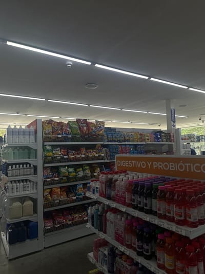 The image depicts the interior of a grocery store, presumably located in Parroquia Ocumare, Venezuela, given the presence of Spanish-language signage. The focus is on the aisles, which are lined with shelves stocked with a variety of snack foods and beverages.
Prominently featured is a sign advertising "DIGESTIVO Y PROBIÓTICO," suggesting a section dedicated to digestive aids and probiotic products, consisting of drinks. Adjacent to this, shelves are filled with various brands and flavors of chips, displayed in their packaging.
Below the chips, a lower section houses larger containers of liquids and other products, possibly cleaning supplies or larger format beverages. Fluorescent lighting fixtures are visible on the ceiling, providing illumination to the store. The scene conveys an everyday shopping environment within a typical grocery store setting.