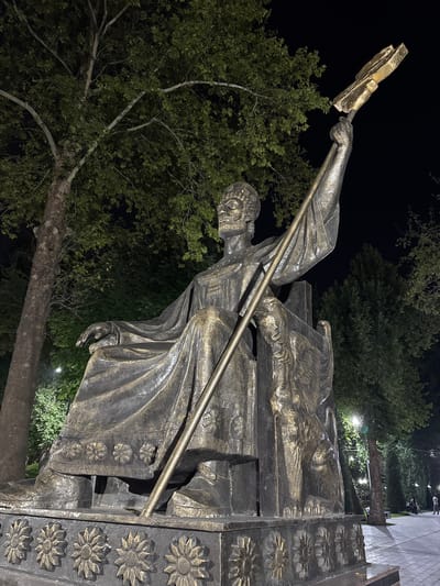 The image shows a bronze statue of Amir Temur sitting on a throne and holding a scepter. The statue is located in Dushanbe, Tajikistan, in a park that is illuminated by streetlights at night. The statue is detailed, with intricate patterns on the throne and the figure's clothing. A lion figure is visible on the side of the throne. Trees with dense foliage surround the statue, and in the background, there are more trees, benches, and a paved walkway where a few people are walking or sitting. The sky is dark, indicating it is nighttime.
