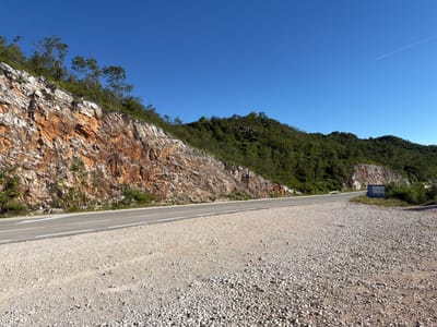 A clear, sunny day is depicted in this outdoor shot, which appears to be near the city of Ponikvica, Montenegro. In the foreground, a dirt and gravel shoulder lines a two-lane asphalt road. White lines mark the lanes, with an arrow visible on the left lane. To the left of the road, a steep, rocky incline is covered in patches of green vegetation and a protective wire mesh. The rock face exhibits striking orange and brown striations. Beyond the incline, a lush, green forest blankets the rolling hills. The sky is a vibrant, cloudless blue, with a faint white contrail from an airplane visible in the upper right quadrant. On the right side of the road, a blue information kiosk stands near a dense cluster of trees.