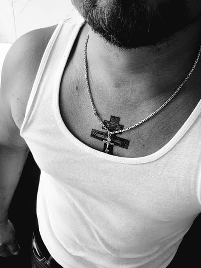 The black and white media file shows a close-up, high-angle shot of a person's chest and neck area, wearing a white tank top. The person has dark hair and a beard visible on the left side of the frame. A silver chain necklace rests on their skin, with a crucifix pendant hanging over a tattoo of a cross. The tattoo appears to be a traditional Orthodox cross. The setting appears to be indoors, with a light-colored, possibly tiled floor visible in the background on the left. The lighting is high contrast, creating shadows and highlighting the textures of the skin, hair, and fabric. There is no visible text in the image. The overall mood is intimate and personal, focusing on religious symbolism and personal adornment.
