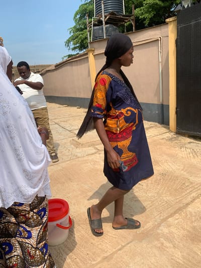 In Shagamu, Nigeria, a pregnant woman wearing a black headscarf and a blue and orange patterned dress walks alongside other individuals. The woman is in profile, facing right, with her hands visible. To her left, a person in a white hijab and a patterned skirt stands partially in view. Behind them, a man in a white polo shirt and brown pants looks down at his phone. The scene unfolds on a paved outdoor area with a textured concrete ground. In the background, a beige wall with a yellow pillar and a dark metal gate can be seen. Overhead, the sky is clear and blue, with the tops of green trees visible above the wall. A black water tank is mounted on a structure to the left. A red-lidded bucket is placed on the ground near the individuals.
