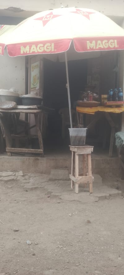 The image depicts an outdoor shop entrance situated on unpaved ground in the city of Muhammadu Dikko Stadium, Nigeria. A prominent cream-colored umbrella, branded with "MAGGI" in red text and a red star logo, partially extends over the shop's doorway, providing shade. The entrance itself is a dark, open void in a light-colored building.
In the foreground, slightly to the right of center, a light-toned, unvarnished wooden stool supports a clear plastic bucket containing a dark, opaque liquid. The ground immediately surrounding the stool and leading up to the shop is a mix of packed dirt and small gravel, with cracked, irregularly shaped concrete pavers directly in front of the building's raised threshold.
To the left of the entrance, two rustic wooden tables are visible. The front table holds at least two large, lidded metal containers and a small, shallow, woven basket. To the right of the entrance, another wooden table, draped with a crumpled yellow fabric, displays merchandise including several clear plastic bottles of water, packaged items (likely snacks) stacked in a woven basket, a small red cylindrical object, and a cardboard box partially inscribed with "Woman is a Star." No individuals are visible in the scene, and no explicit actions or interactions are depicted.