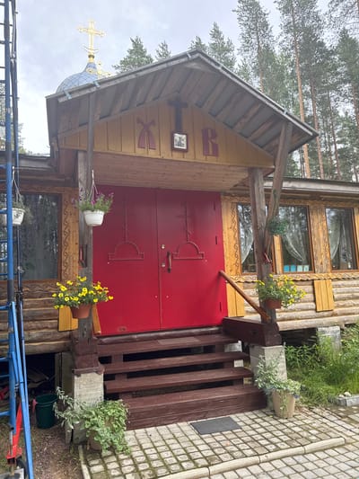 The image shows the exterior of a wooden church building in Nenyalki, Russia. The church has a red double door entrance with crosses on each door. Above the entrance, there is a wooden roof and a smaller decorative roof section with Cyrillic letters and a religious icon. The building is constructed of logs and has wooden window frames. Hanging flower baskets are placed at the entrance. The church is surrounded by trees, suggesting a rural or semi-rural setting. Scaffolding is visible on the left side of the image. The ground in front of the church is paved with cobblestones, and there are potted plants and grass in the foreground. The sky is overcast.