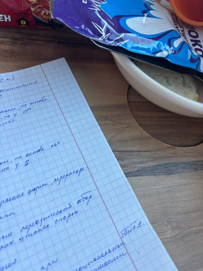 This scene, located in Chany, Russia, depicts a close-up, high-angle view of a wooden surface. In the lower left and central foreground, a white, grid-lined notebook featuring blue squares and a red vertical margin line displays blue handwritten text in Russian. In the upper right quadrant, a white ceramic bowl contains a piece of white bread. Resting on the rim or within the bowl, a blue and white plastic package is visible, partially obscured, with visible text including 'МОЛОКО СГУЩЕННОЕ С САХАРОМ' (CONDENSED MILK WITH SUGAR). An orange cylindrical object, possibly a cup, is partially visible adjacent to the blue and white package. In the upper left quadrant, a red plastic package is partially visible, displaying 'ОТЛИЧНЫЙ ВКУС' (EXCELLENT TASTE) and the number '5'. No individuals are present in the image.