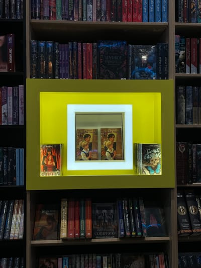 This is a medium-shot, eye-level interior photograph of a bookstore shelf in the city of Kommunarka, Russia. The shelves are filled with books. In the center of the image, a prominent yellow display frame with a white inner border is featured. Inside the frame, two books are visible, seemingly displayed behind glass. To the right of this display, a single book stands upright. The surrounding shelves are densely packed with various titles. The overall environment suggests a retail setting dedicated to the sale of books. There are no people visible in the photograph.