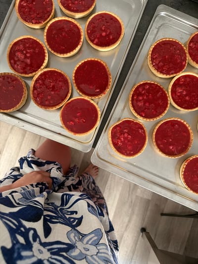 The image depicts a close-up, overhead view of two metal baking trays filled with small, round fruit tarts topped with a vibrant red fruit filling. The tarts have a golden-brown crust and appear to be arranged in rows. In the foreground, a person's legs and lower torso are visible, clad in a blue and white floral patterned garment. The person's feet are also visible, one of which is slightly curled. The baking trays are resting on a light-colored, wood-planked floor. The scene is likely indoors, possibly in a kitchen or food preparation area. The specific location in Meisenheim, Germany, is not discernible from the image itself.