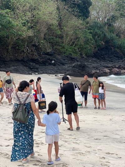 The image depicts a beach scene in Manggis, Indonesia. A group of people, including adults and children, are walking on a sandy beach with lush green vegetation and dark volcanic rocks forming a backdrop. Some individuals are carrying bags, and one person appears to be setting up camera equipment. The sea with gentle waves is visible in the background, and the overall atmosphere suggests a casual outing.