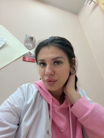 The image shows a young woman taking a selfie in what appears to be an indoor setting with light pink walls. She is wearing a pink hooded sweatshirt underneath a white jacket. Her hair is dark and pulled back. She is wearing earrings and has a hand near her neck. There are some papers or posters visible on the wall behind her, one with text in Russian. The overall impression is a casual, personal photograph. The location specified, Гаревая, Russia, is not visually supported by the image content.