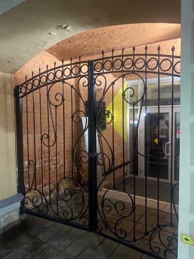 The image shows a closed black wrought iron gate in front of a glass-paneled door. The gate features decorative scrollwork and vertical bars. Behind the gate, the glass door is visible, with a reflection of a person in a suit and tie. A yellow sign with a black arrow pointing down is attached to the wall above and to the right of the door. The background shows a brick wall with a yellow and brown patterned area, and a concrete floor. A peeling section of a light-colored wall is visible on the left side of the image, with a textured orange brick wall behind the gate. The scene appears to be an entrance to a building or a courtyard. The lighting suggests it might be evening or indoors with artificial lighting. There is a sign with Cyrillic text above the door, though it is mostly obscured by the gate. A smaller blue and red sign is also visible on the door. The overall impression is one of an enclosed, perhaps historical, urban setting.