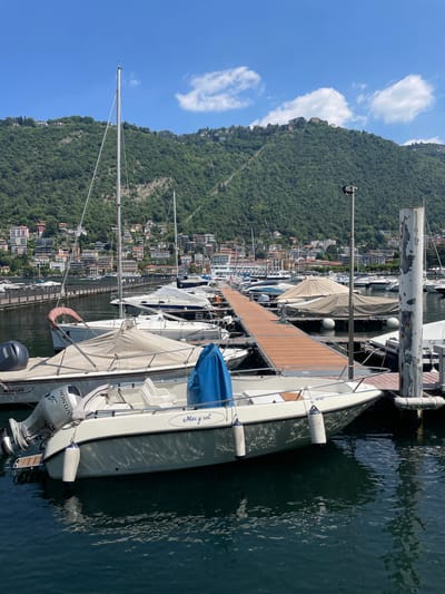 The image shows a marina on Lake Como, Italy. The location is recognizable by the lush green mountains rising behind a townscape with terracotta rooftops and buildings. The foreground features several boats docked side-by-side along a wooden pier. The boats are of various sizes and types, including sailboats and motorboats. A white sailboat with the name "Marysol" is closest to the viewer. The water in the marina is dark and slightly reflective. The sky is blue with a few scattered clouds.