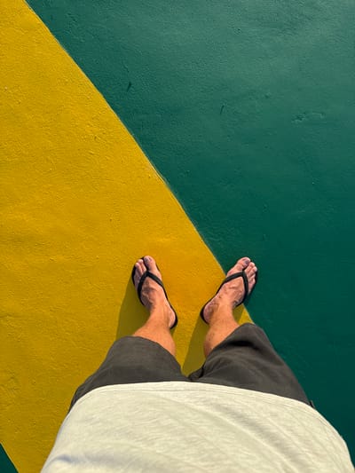A high-angle shot depicts a person standing on a surface divided diagonally into green and yellow sections. The person is wearing black flip-flops, dark gray shorts, and a white shirt that is partially visible. The surface texture appears to be rough. The location is inferred to be Pasir Panjang, Indonesia.