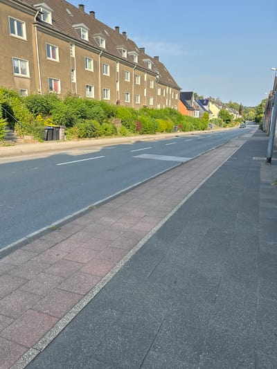 The image depicts a street scene in Velbert, Germany, specifically on Langenberger Straße. The perspective is from the sidewalk, looking down the street. On the left side of the street, a row of multi-story residential buildings with pitched roofs and numerous windows is visible. These buildings are bordered by dense green shrubbery. Further down the street, other houses and trees are visible, indicating a residential area. The street itself is paved asphalt with white lane markings. A sidewalk runs along the right side of the street, composed of two sections: an inner section with red pavers and an outer section with dark gray pavers. Two black trash bins are positioned on the left side of the road, near the residential buildings. A few cars are visible driving on the street in the distance. The sky is clear and blue, suggesting fair weather.