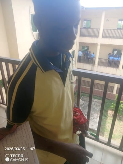 The scene is an outdoor setting in Nsugbe, Nigeria. A young individual, partially silhouetted and visible from the chest up, occupies the foreground, looking downwards and to the right. This person wears a yellow polo shirt with navy blue sleeves featuring yellow stripes and a visible emblem on the chest. A thin silver-colored chain is present around the neck. The right hand holds a green pen and a crumpled red fabric object, while the left hand holds a piece of paper displaying handwritten text and a '2026/03/05 11:07 TECNO CAMON 19' timestamp. The individual is positioned behind a brown metal balcony railing.

In the background, a multi-story building with a light-colored facade and matching brown balcony railings is observed across an open space. On its lower balcony, a group of approximately 10-15 individuals, identified as students by their light blue uniforms, are standing and conversing. Additional individuals are visible on the building's upper balcony. The ground area below the background building consists of concrete or pavement with minimal green vegetation. The lighting indicates daytime.