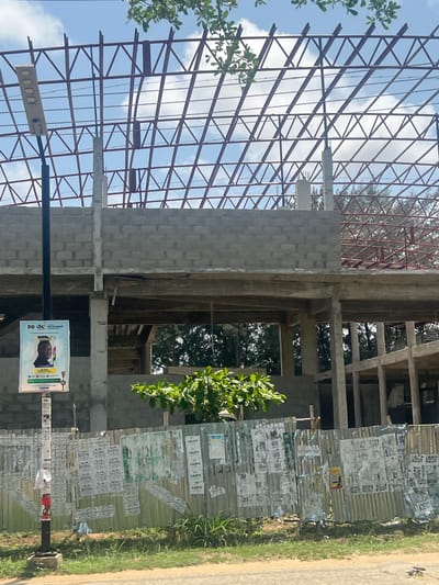 The image depicts a construction site in Ijanikin, Nigeria. An unfinished multi-story building stands prominently, its skeleton of concrete blocks and steel framework exposed. Scaffolding and support structures are visible within the building's open spaces. A metal fence, covered with posters and advertisements ("RUPAL", "KAPPO", "JIGGY JISGY"), runs alongside the construction site. A street light pole with a political advertisement attached is in the foreground. Vegetation is visible on the periphery of the scene, including a small tree in front of the building and grass along the side of the road. The sky is partially cloudy.