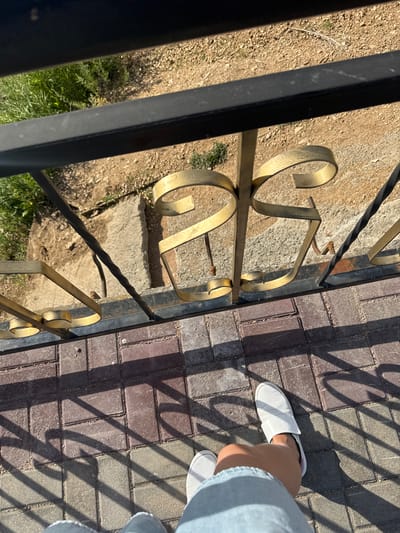 The image shows a close-up, Dutch angle shot of a black wrought iron fence with decorative gold elements. To the right of the fence, a person's legs and feet are visible, wearing white slip-on shoes and light blue denim pants. They are standing on a paved walkway made of rectangular bricks in various shades of red and gray. The sun is casting strong, angular shadows from the fence onto the walkway, indicating it is likely a sunny day. The ground to the left of the fence is rough, with dirt, small stones, and patches of green grass and dry vegetation. The scene appears to be outdoors, possibly in a park or residential area, given the fence and paved walkway. The lighting suggests it is daytime, likely late morning or early afternoon. There is no visible text in the image. The overall impression is a mundane moment captured on a sunny day, focusing on the textures of the fence, pavement, and the person's attire. The location context provided is Kokshetau, Kazakhstan, which does not add specific visual details to the image itself.