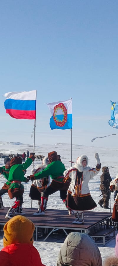 The scene depicts an outdoor cultural performance in Nelmin-Nos, Russia, set against a vast snow-covered landscape under a clear blue sky. On a raised, dark wooden stage, a group of at least five individuals are performing traditional dances. Performers are dressed in intricate indigenous attire, featuring fur trims, patterned boots, and vibrant colors such as green, brown, white, red, and yellow. A female performer, center-right, wears a white fur-hooded coat with colorful braids and is captured mid-movement with her right arm raised. Two male performers to her left wear green tunics over brown trousers, with patterned boots, and are engaged in a synchronized movement. In the midground, two flags fly from poles: the Russian national flag (white, blue, red horizontal stripes) on the left, and the flag of the Nenets Autonomous Okrug (white and blue with a central circular emblem) on the right. Behind the performers and flags, a herd of reindeer is visible on the snowy plains, accompanied by several distant human figures. In the immediate foreground, the backs of two spectators, one in an orange and yellow hooded jacket and another in a grey puffer jacket, are visible, observing the stage. The environment is characterized by extensive snow coverage and distant, low-lying hills.