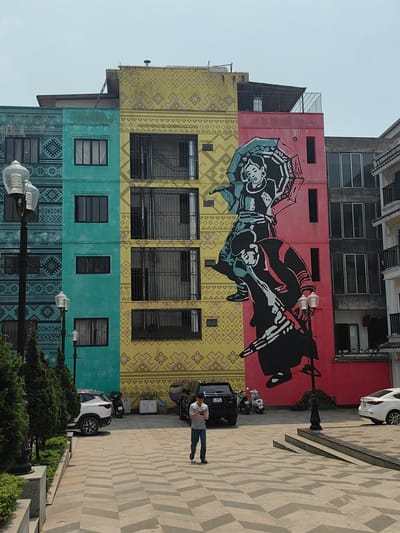 The image displays an urban street scene in Sa Pa, Vietnam, centered on a large building facade. The building is segmented into three vertical sections: a teal-colored section on the left, a yellow section in the middle, and a pink section on the right. Both the teal and yellow sections are adorned with repetitive geometric patterns. A large mural, primarily in black, white, and teal, covers parts of the yellow and pink sections. This mural depicts two stylized figures in traditional attire; the upper figure holds an open umbrella while the lower figure is positioned beneath.
In the foreground, a paved plaza area with a chevron-like pattern in light and dark gray extends from the bottom of the frame towards the building. A single male individual walks towards the viewer from the middle ground, looking down at a white smartphone held in both hands. He is wearing a light-colored, short-sleeved patterned shirt, blue jeans, and dark sandals.
To the left, a series of green conical shrubs are planted along a raised curb, adjacent to a dark lamppost with a clear, multi-faceted glass fixture. Another similar lamppost is visible further back. Vehicles parked near the building include a white SUV on the left, a black SUV, a white scooter behind the black SUV, and a partially visible white sedan on the far right. The sky is clear and bright, indicating daytime conditions.