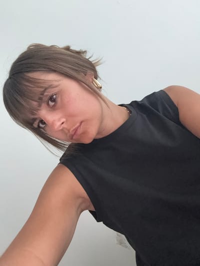 A woman is seen from a low angle, her head tilted toward the camera. She has light brown hair with bangs, brown eyes, and fair skin. She wears a black t-shirt and gold earrings. The background is a plain white wall. There is no information about the location in Cannes, France, or any other city.
